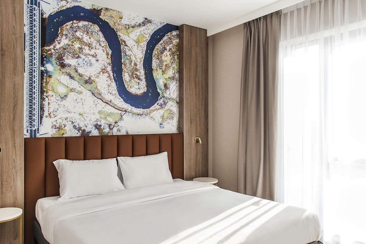 Une chambre d’hôtel avec un grand lit, une tête de lit, une œuvre d’art murale, une fenêtre avec des rideaux et une petite table de chevet.