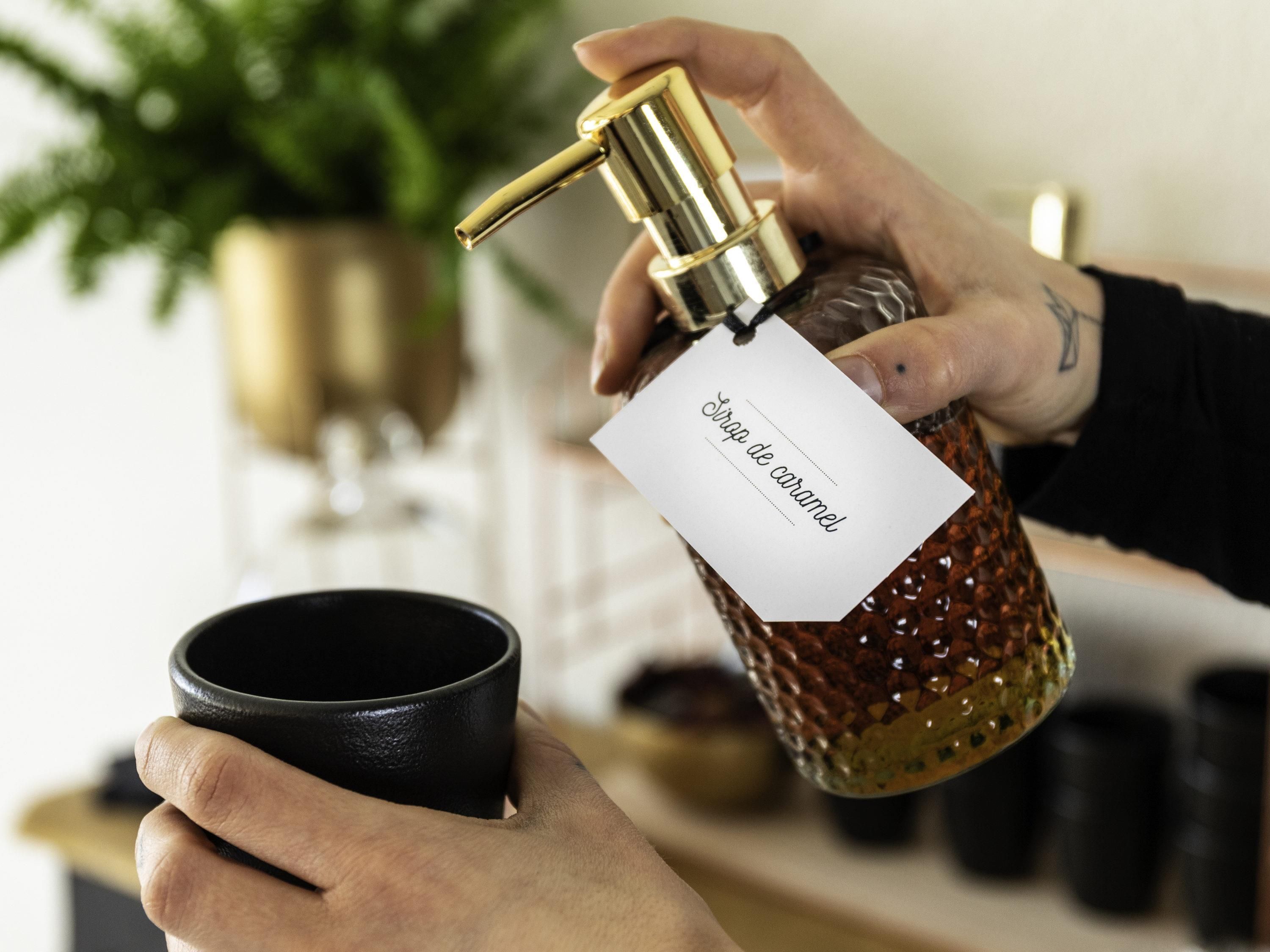 Une main tenant une bouteille dont l’étiquette indique « sirop de canne à sucre » et une tasse noire.