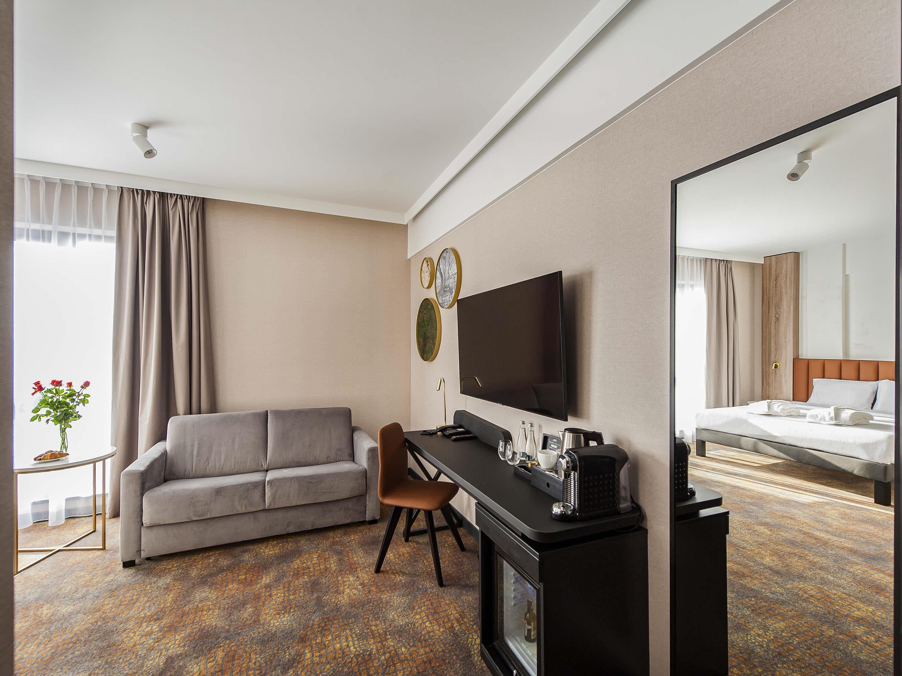 A hotel room with a sofa, a desk with a flat-screen TV, a bed, and a mirror.