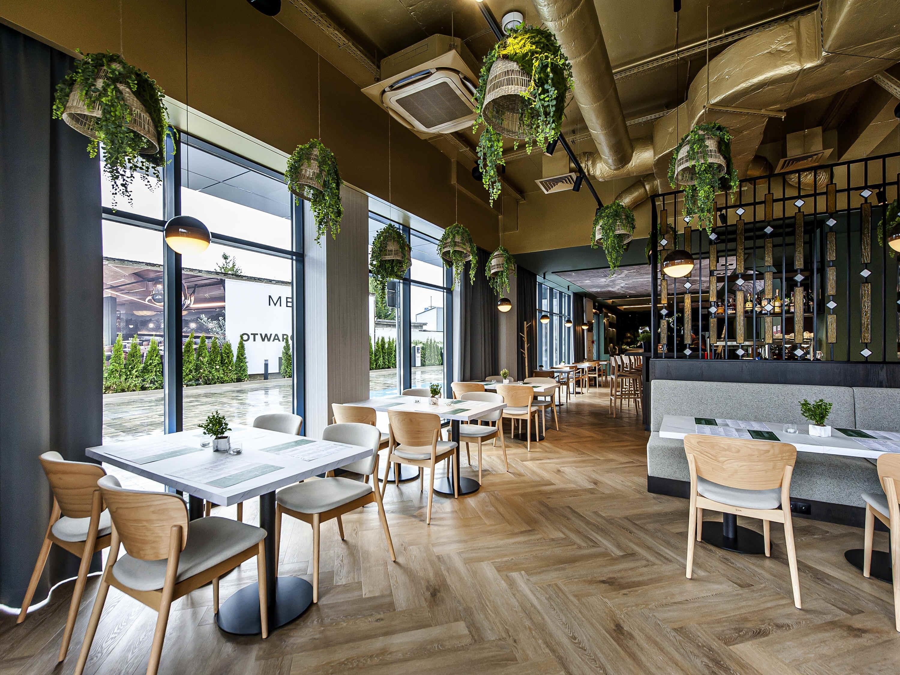 A restaurant interior with wooden flooring, tables, chairs, hanging plants, and large windows.