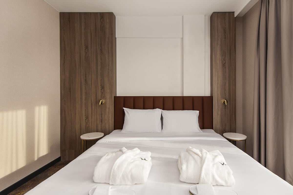 Une chambre d’hôtel avec un grand lit, deux tables de chevet, une tête de lit en bois et une fenêtre avec des rideaux.