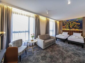A hotel room with a sofa, two beds, a small table with a vase, and a large painting on the wall.