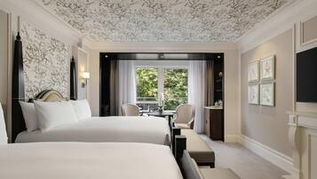 A hotel room with two beds, a dining area, a view of greenery, and framed artwork on the wall.