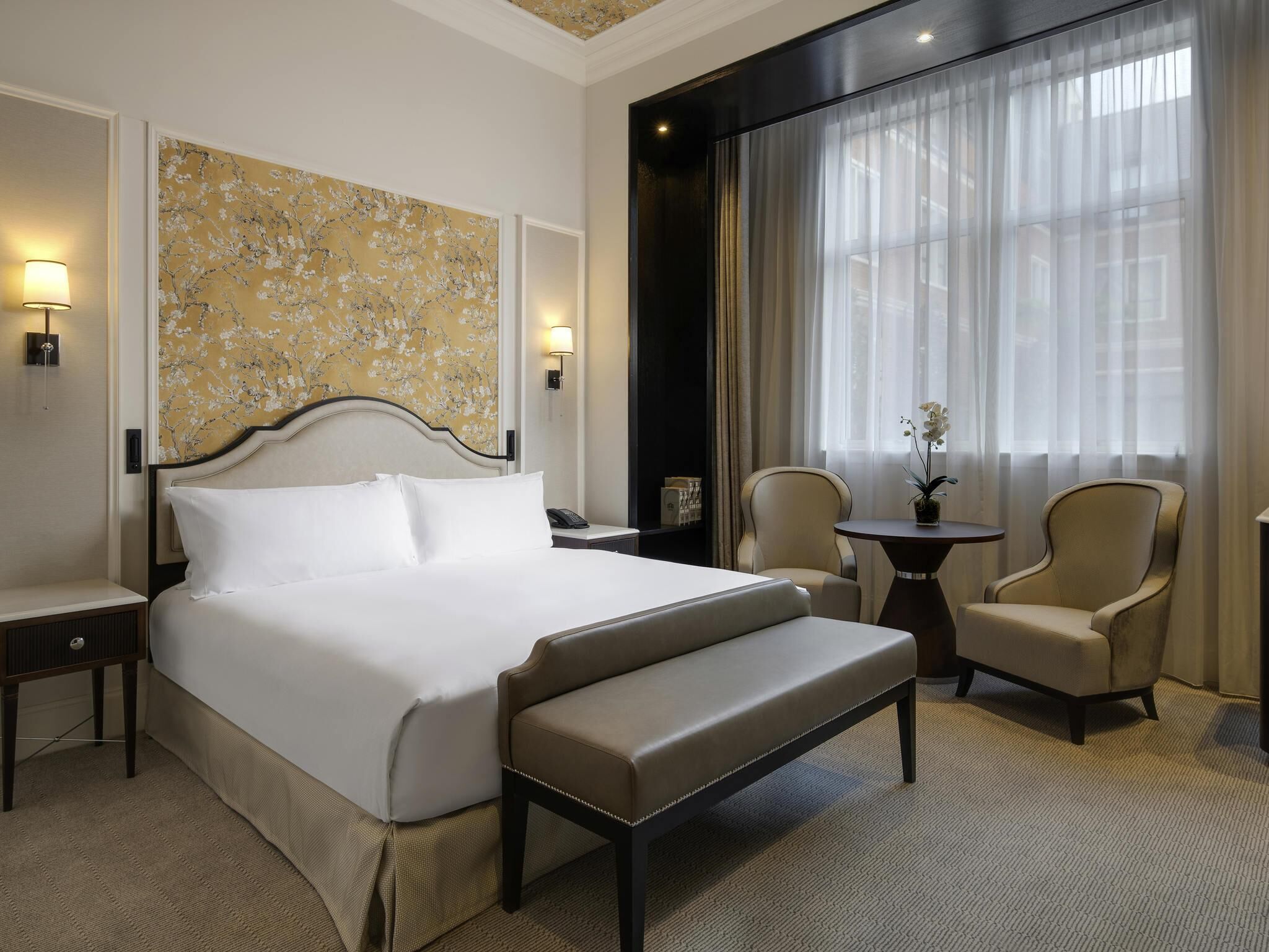 A hotel room with a large bed, two armchairs, a small table, and a large window with sheer curtains.