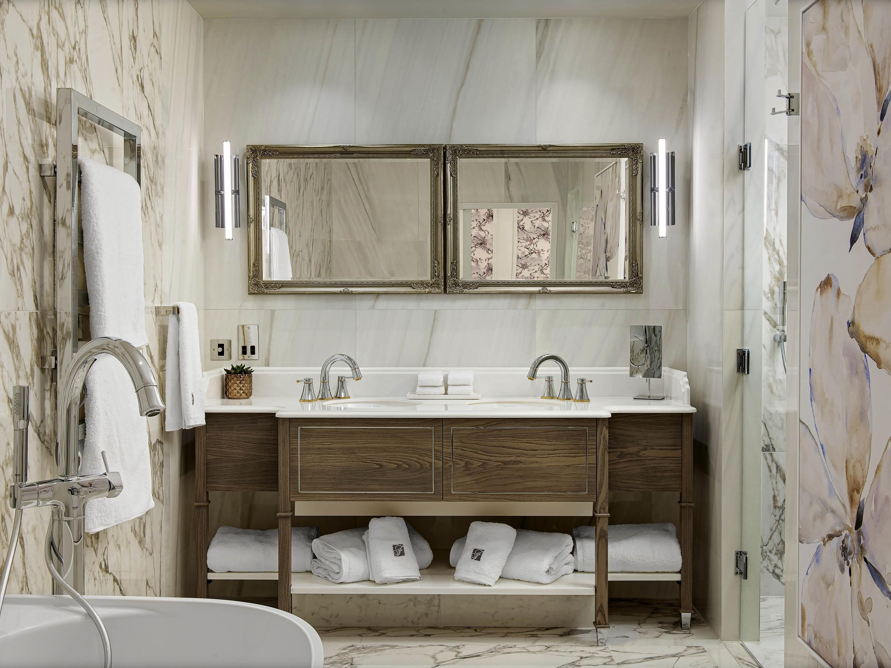 Un baño con pared de mármol, tocador doble con dos lavamanos, un espejo grande y zona de ducha.