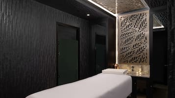 Une salle de spa moderne, dotée d’une table de massage, de lambris en bois foncé et d’un mur décoratif orné de motifs de feuilles.