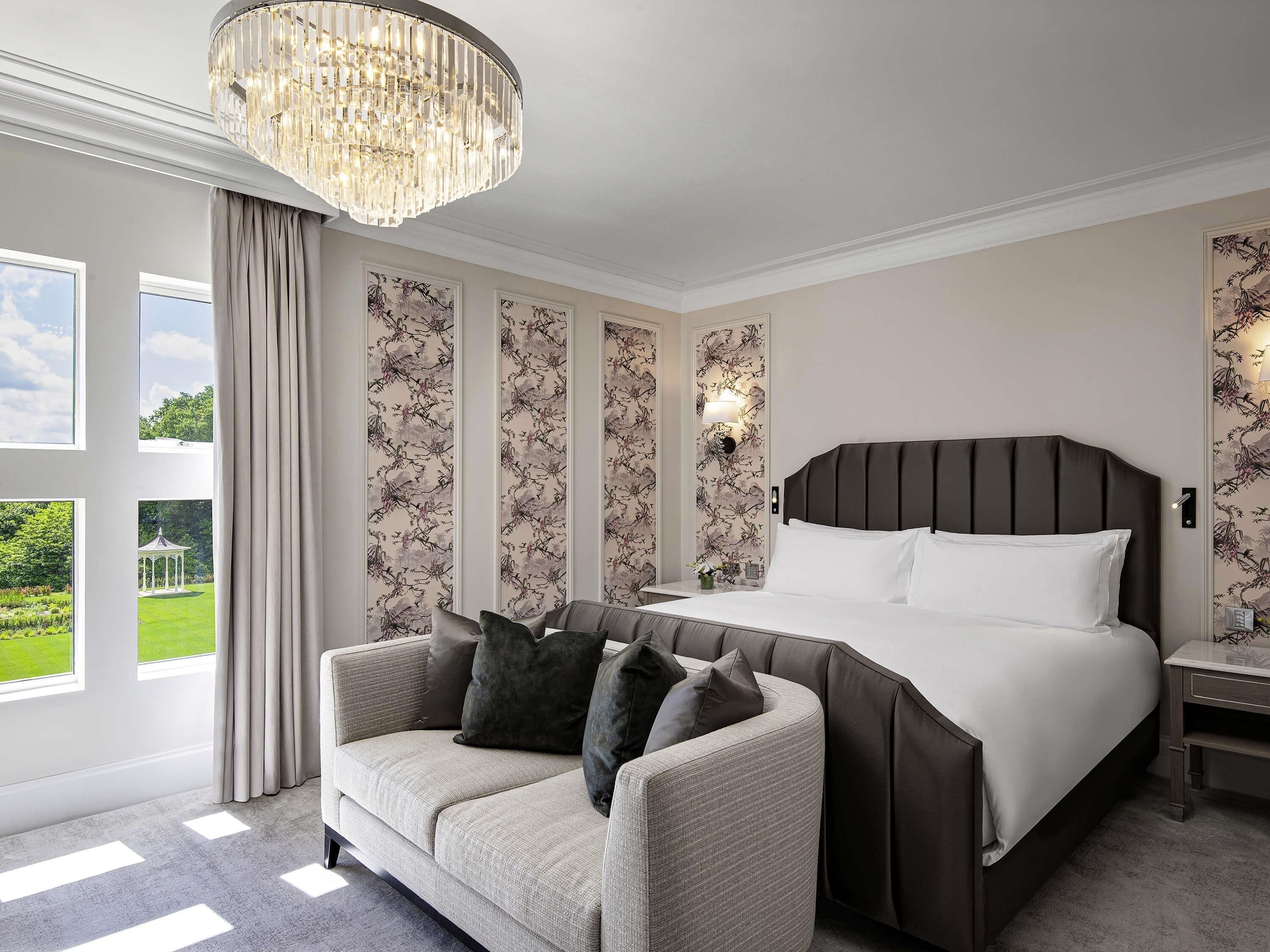 A bedroom with a large bed, a sofa, a chandelier, and a view of a garden.
