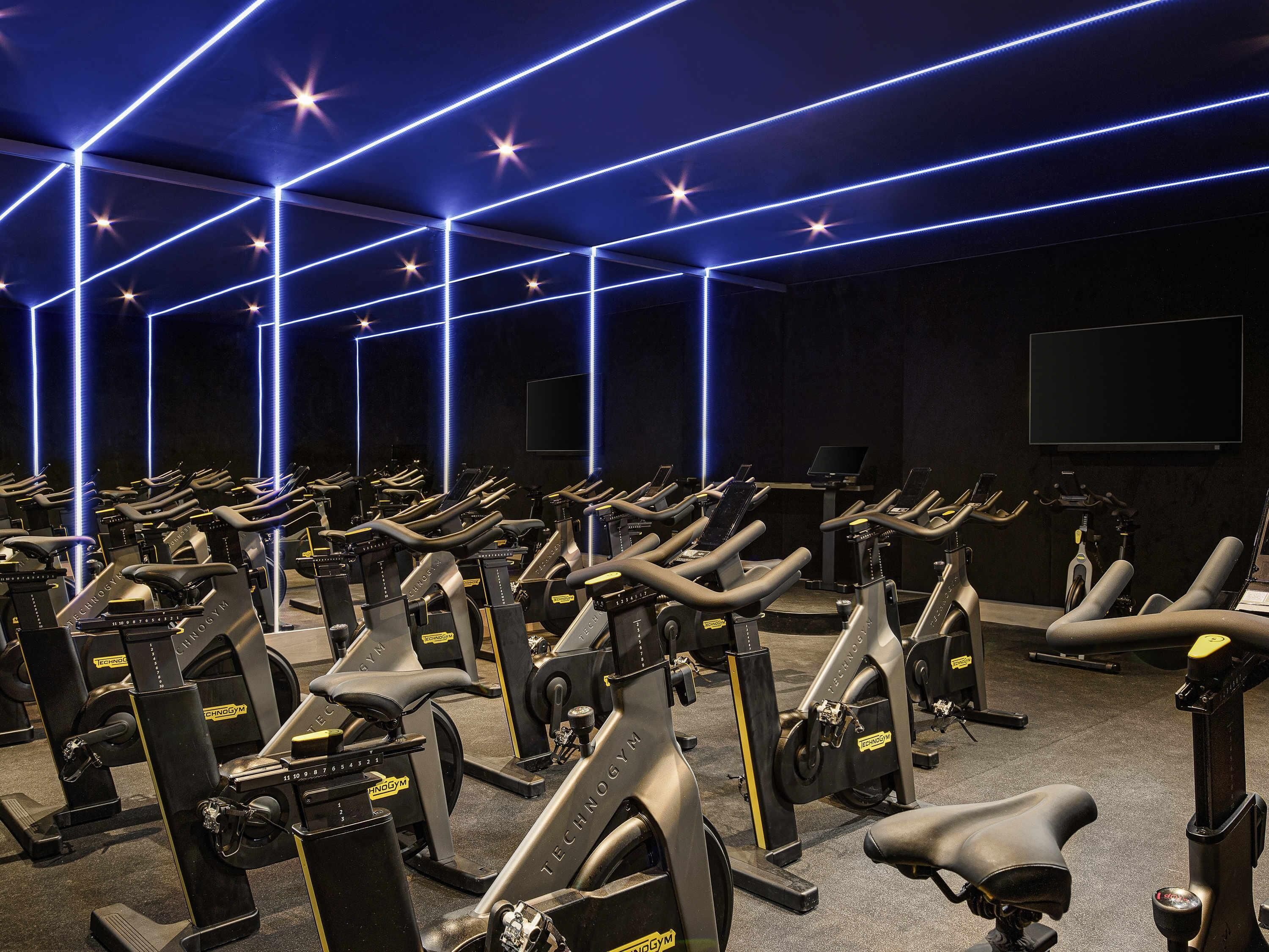 A gym with multiple stationary bikes arranged in rows, a large screen on the wall, and blue LED lighting on the ceiling.