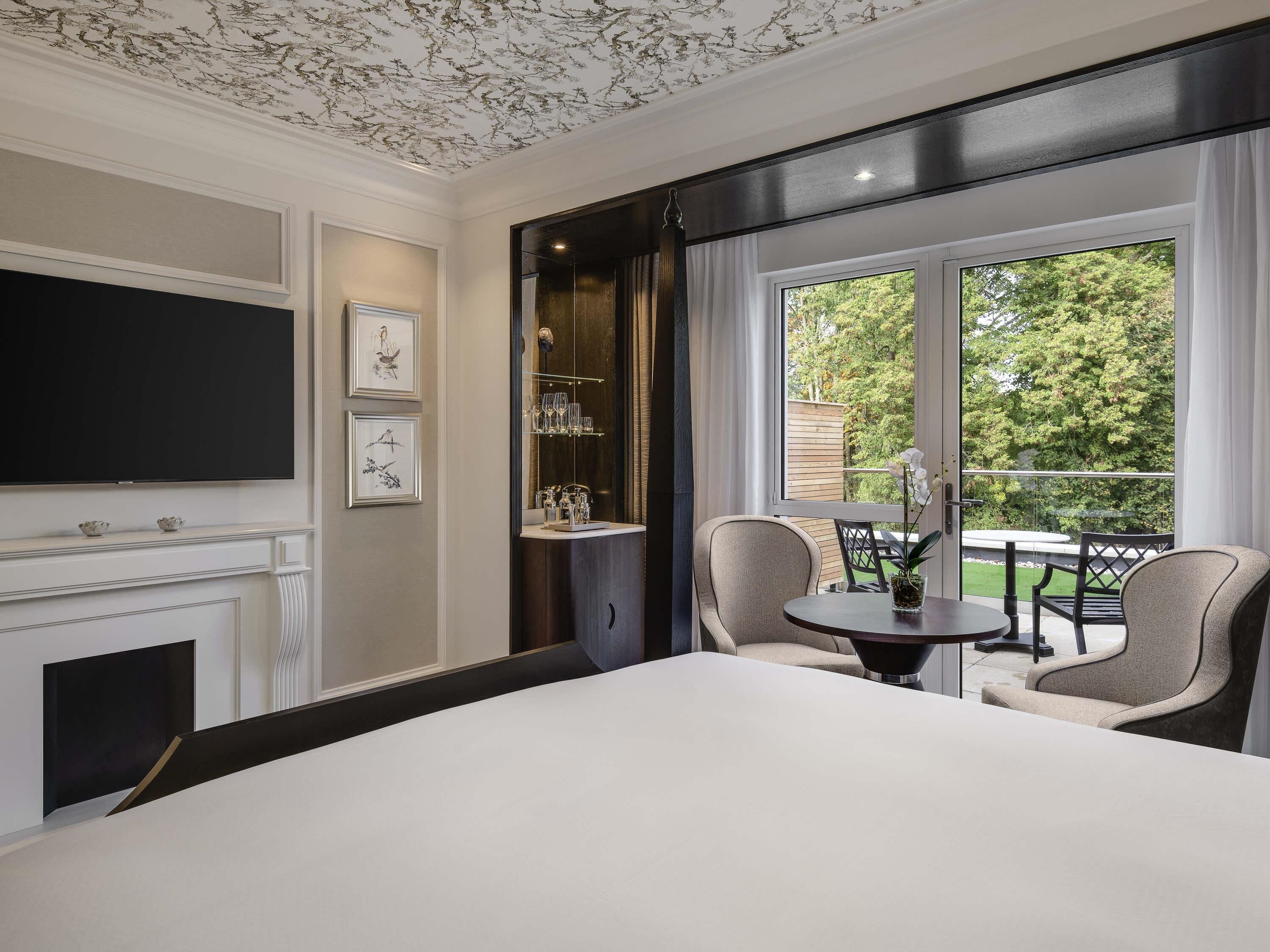 A room with a fireplace, a television, a dining area with chairs and a table, and a window with a view of greenery.