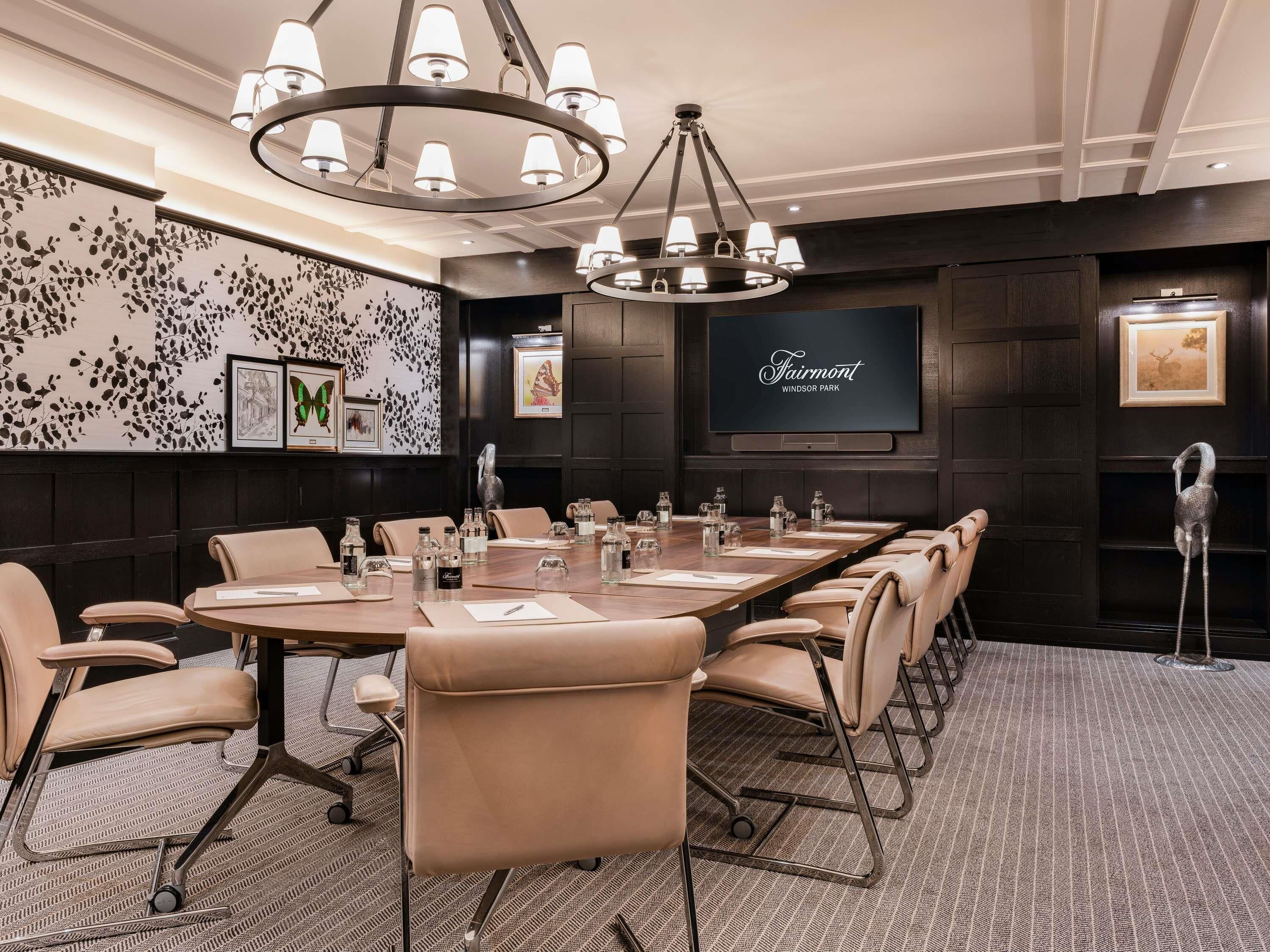 A conference room with a long table, beige chairs, a chandelier, and a screen displaying 'Fairmont Windsor Park'.