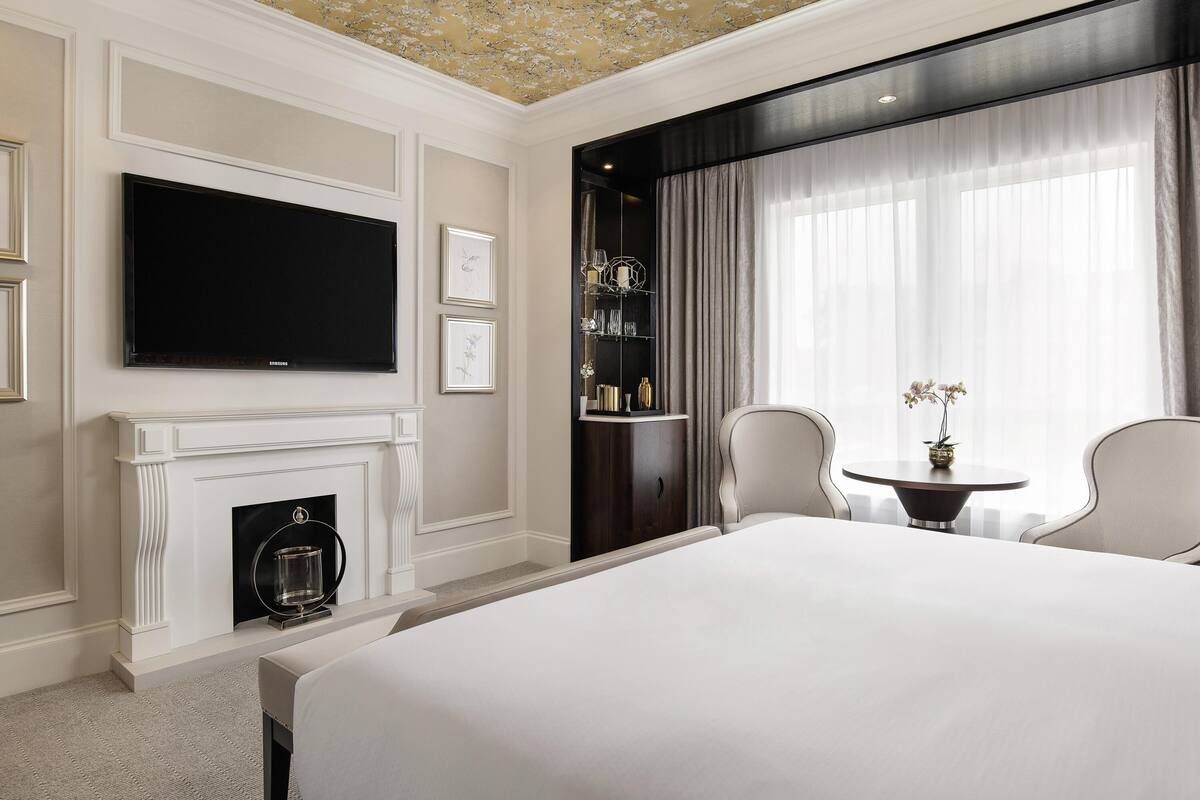 Habitación de hotel con una cama grande, televisor de pantalla plana, chimenea, estantería y vista a la ciudad a través de amplios ventanales.