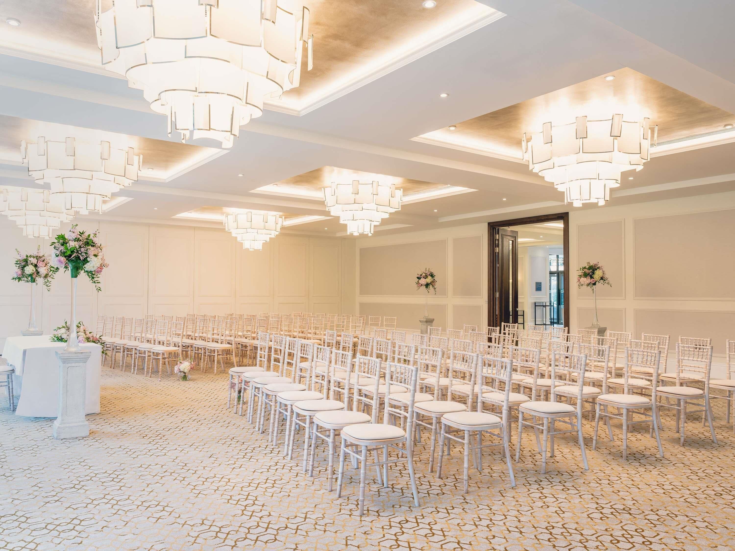 A spacious hall with rows of white chairs, a chandelier, and a large door.
