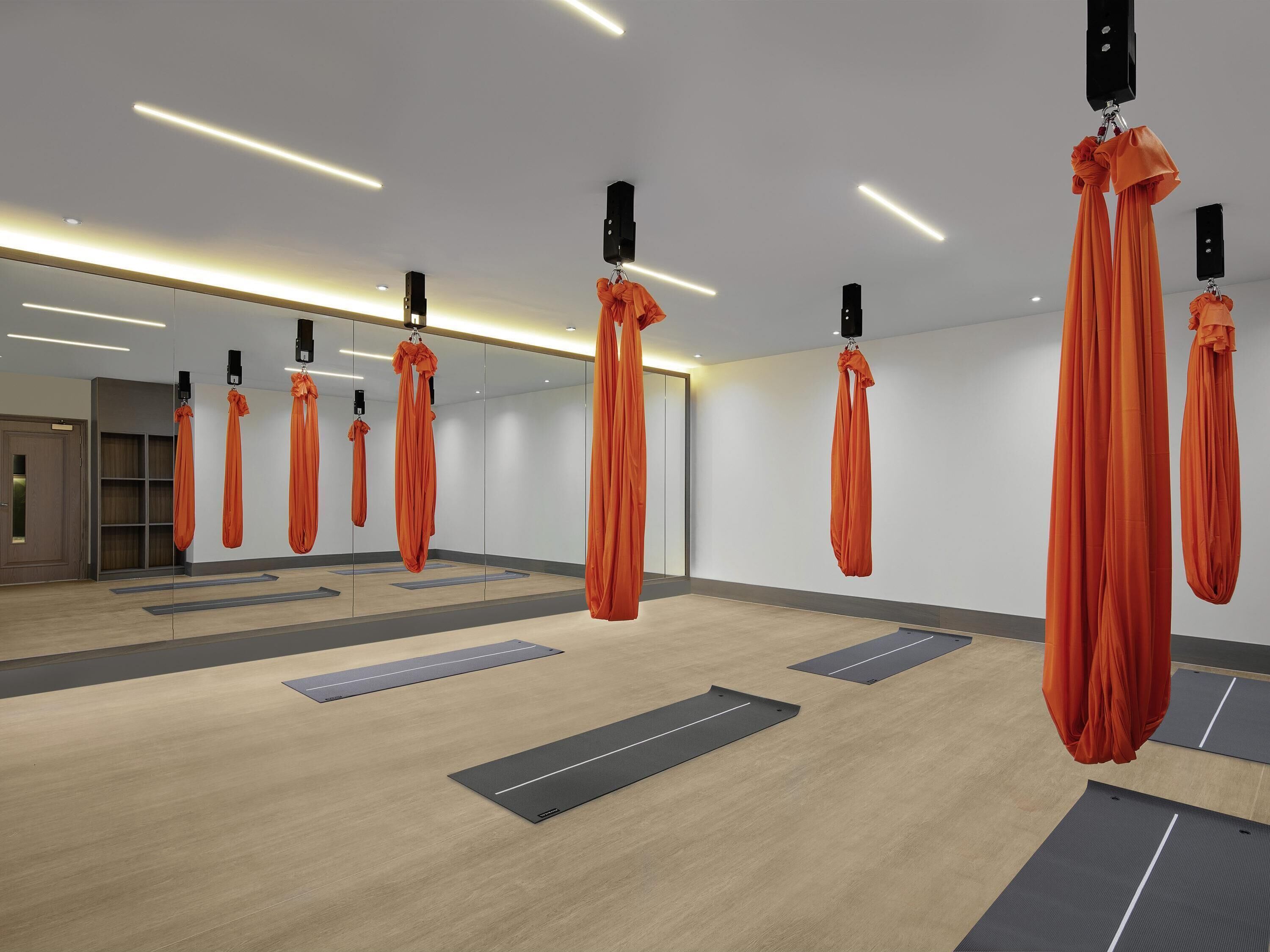 A modern fitness studio with suspended orange aerial silks, wooden floors, and mirrored walls.