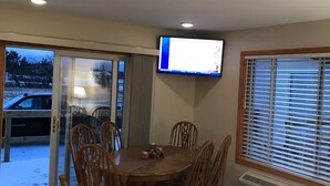Un comedor con mesa y sillas de madera, un televisor montado en la pared y vistas a un espacio exterior con un coche aparcado a través de una puerta de cristal.