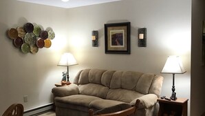 Una sala de estar con un sofá color beige, una mecedora de madera, un tapete estampado y decoración de pared que incluye un cuadro enmarcado y una lámpara de pared.