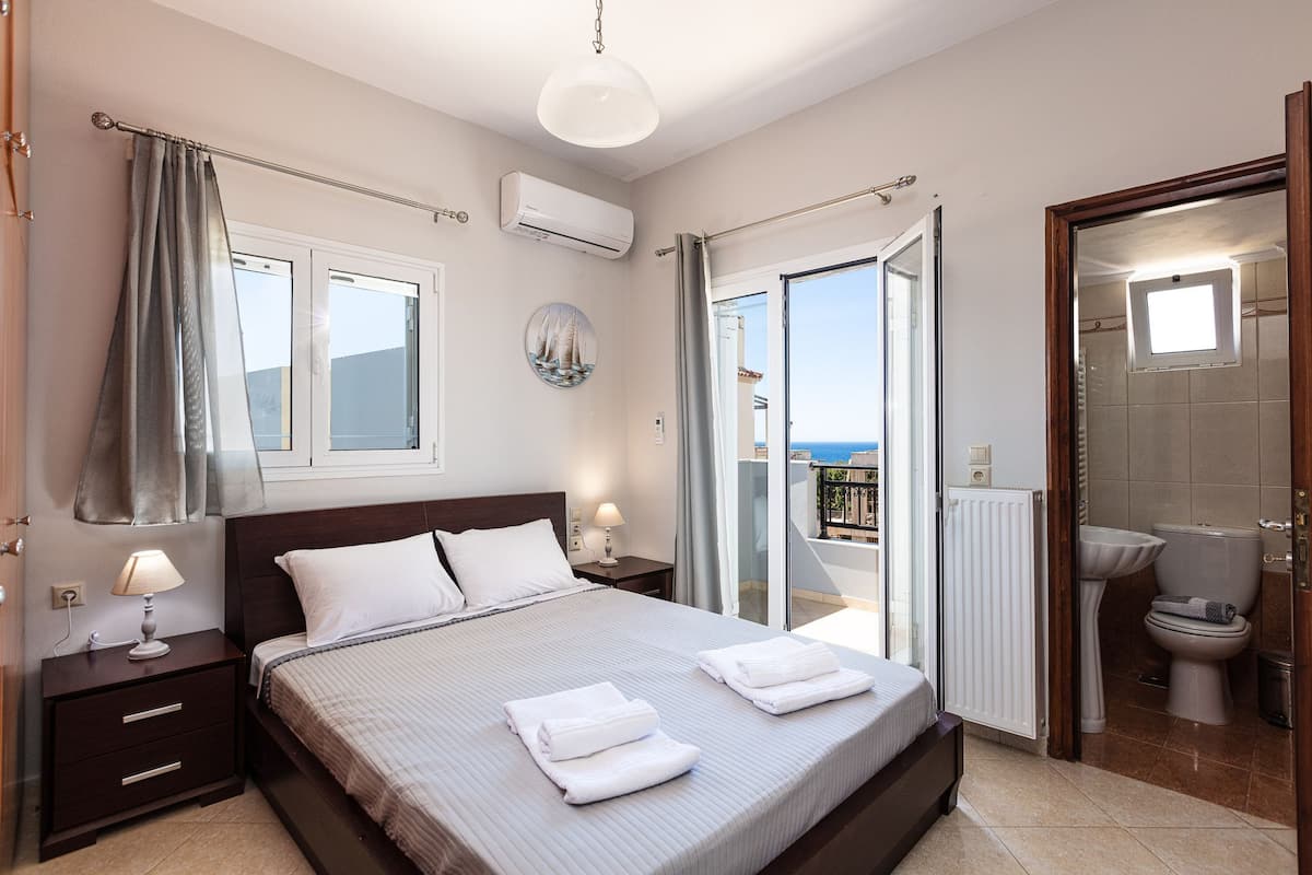 A bedroom with a bed, bedside tables, a window, an air conditioner, and a view of the outside.