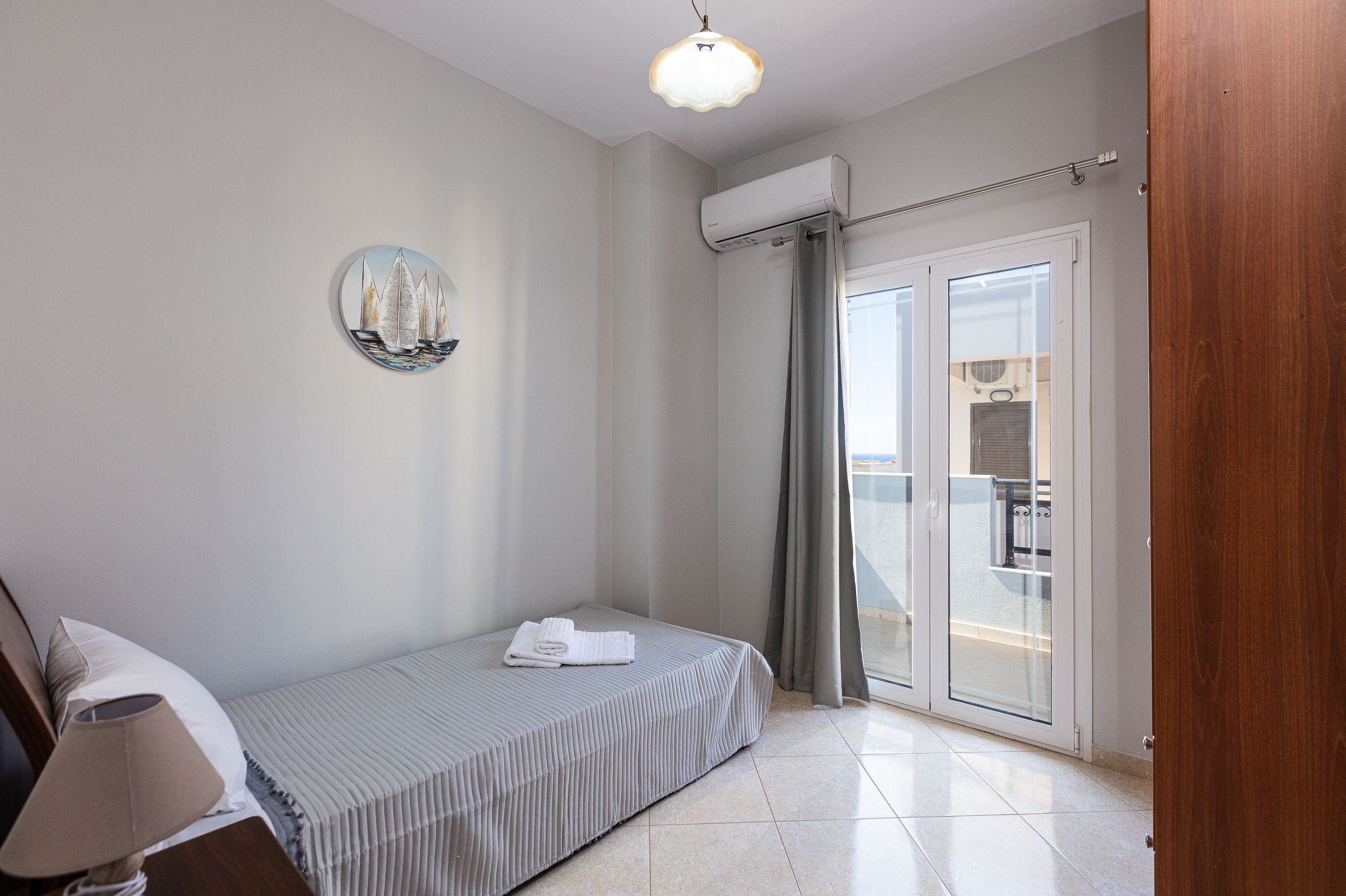 Un dormitorio pequeño y ordenado con cama, lámpara de noche, aire acondicionado instalado en la pared y vistas a través de una puerta corrediza de vidrio.