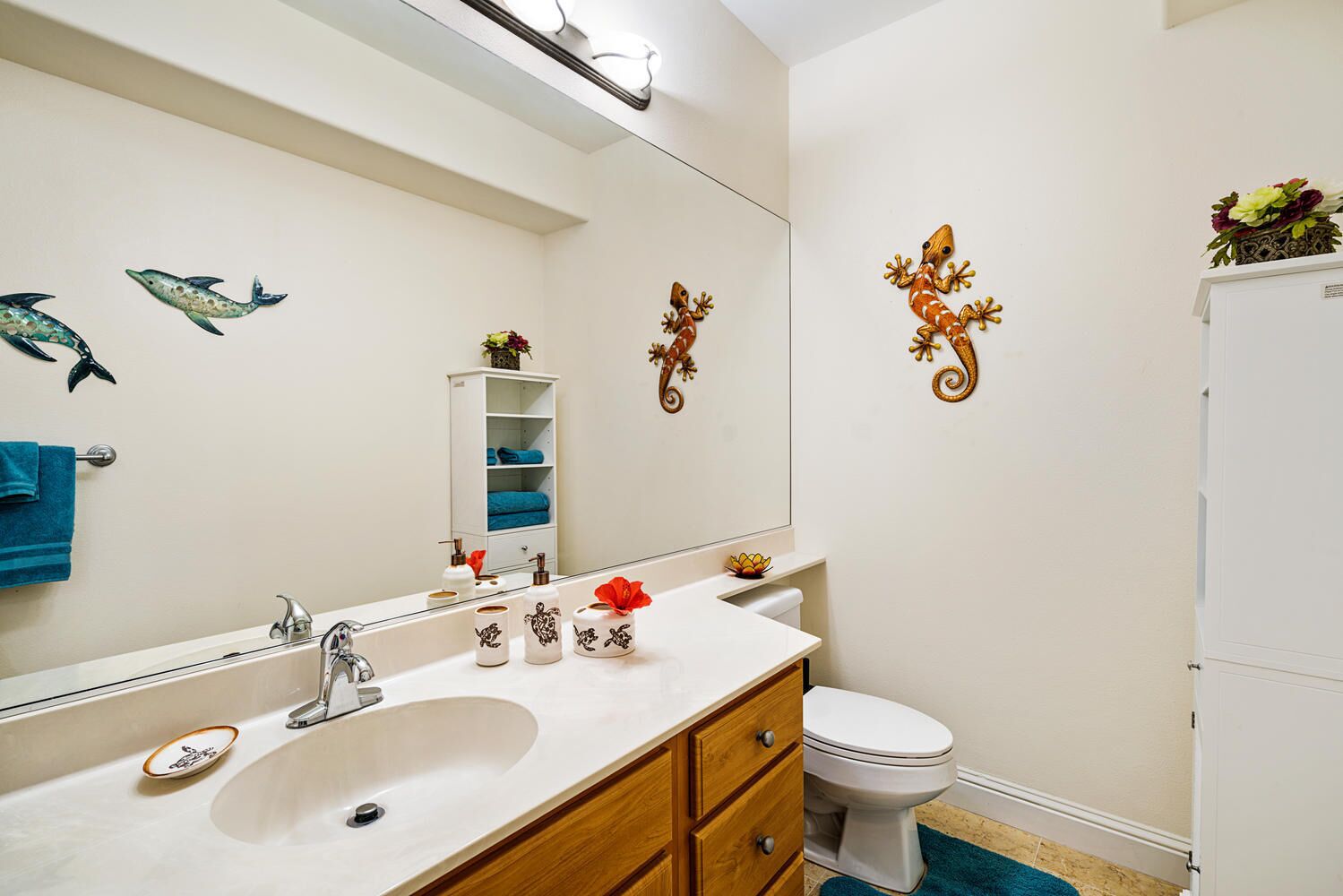 Un baño con lavamanos blanco, tocador de madera, inodoro y detalles decorativos como un espejo, una estantería con toallas y una planta pequeña.