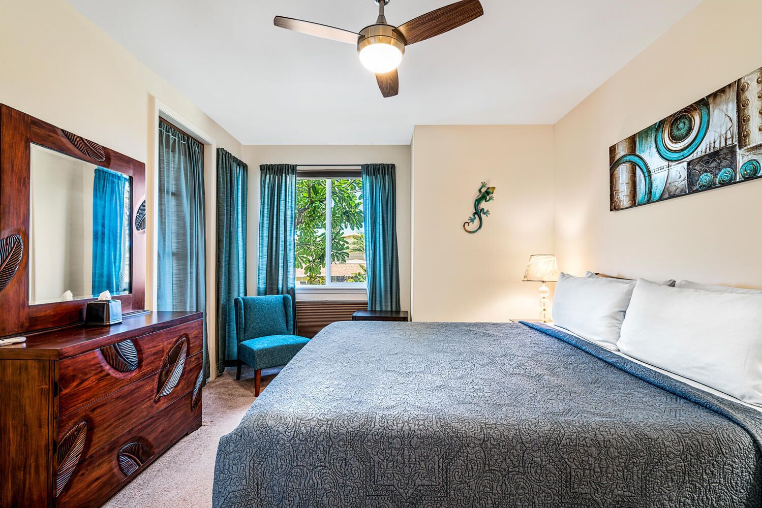 Una habitación de hotel con cama, cómoda, silla, ventilador de techo, ventana con cortinas y un cuadro colgado en la pared.