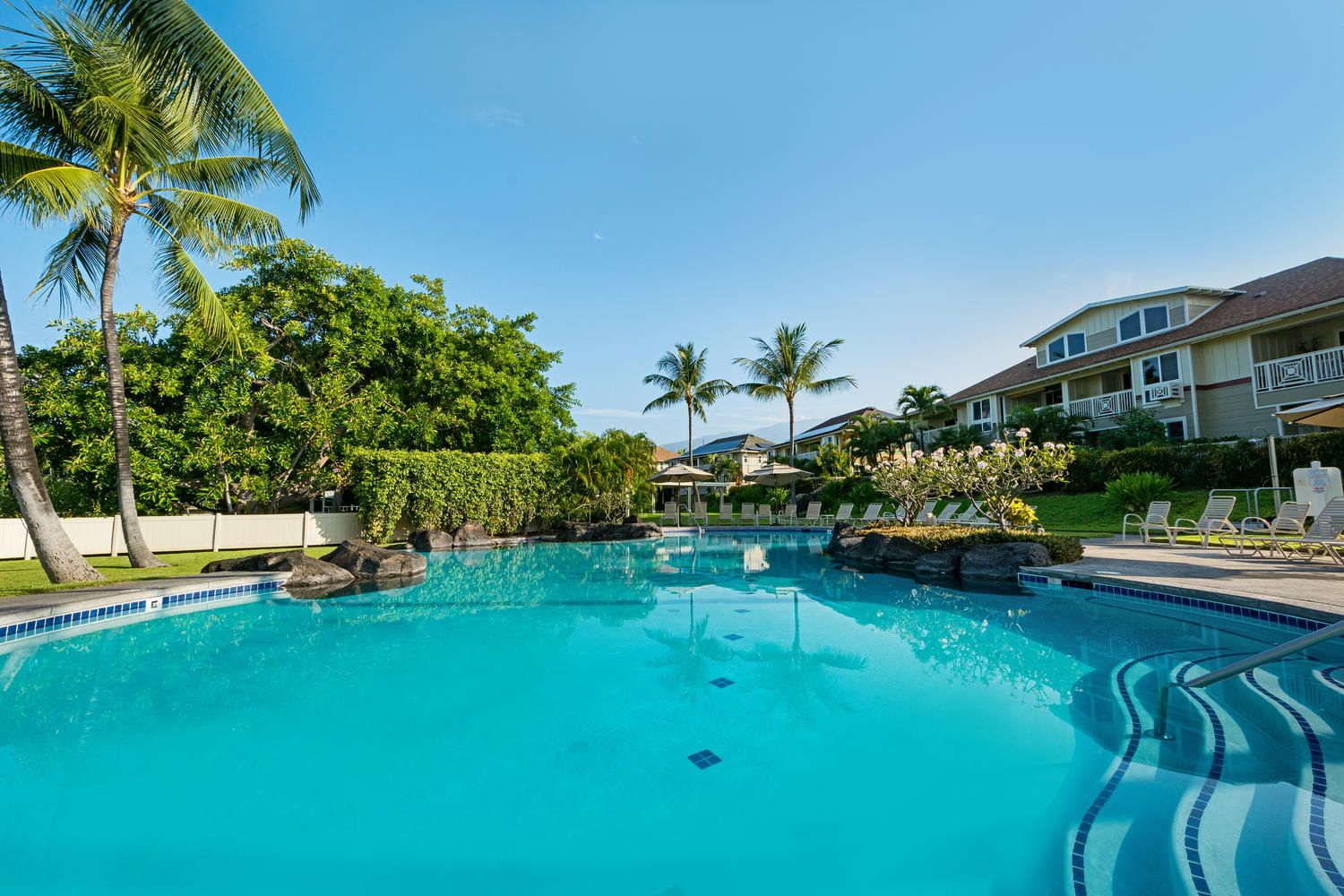 A swimming pool with clear blue water, surrounded by palm trees and a building with balconies.