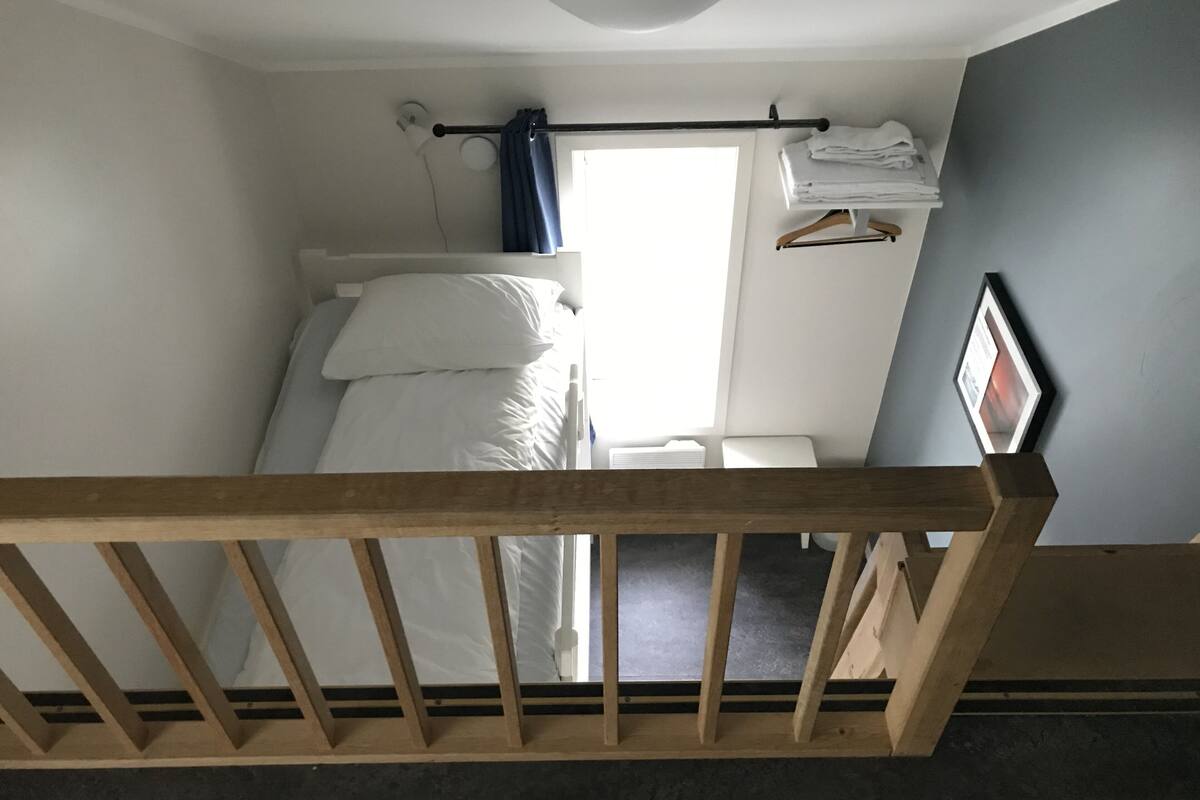 A bedroom with a wooden bed frame, white bedding, a window with curtains, a bedside table, and a wall-mounted heater.