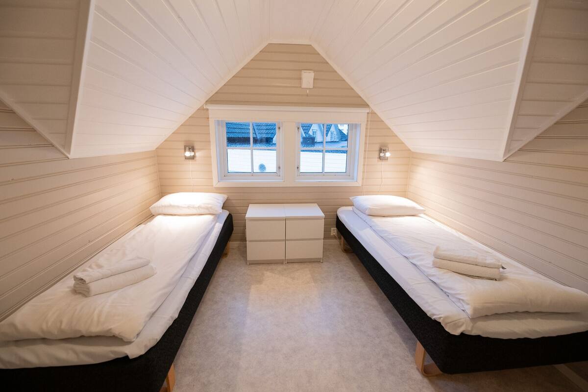 A small, triangular attic bedroom with two single beds, a small table, and a window providing a view of the outside.