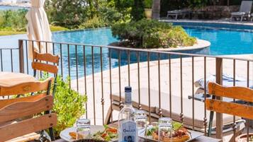 A dining table with a pizza, salad, and drinks, surrounded by wooden chairs and an umbrella, near a pool with a view of the ocean.