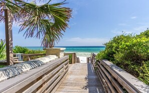 A wooden boardwalk leading to a beach with a palm tree and greenery on the left, and a clear blue ocean on the right.