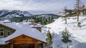 Eine Holzhütte mit verschneitem Dach, umgeben von schneebedeckten Bäumen und Bergen im Hintergrund.