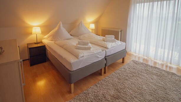 Un lit double avec du linge de lit blanc, deux tables de chevet avec des lampes, un radiateur et une grande fenêtre avec des rideaux légers.