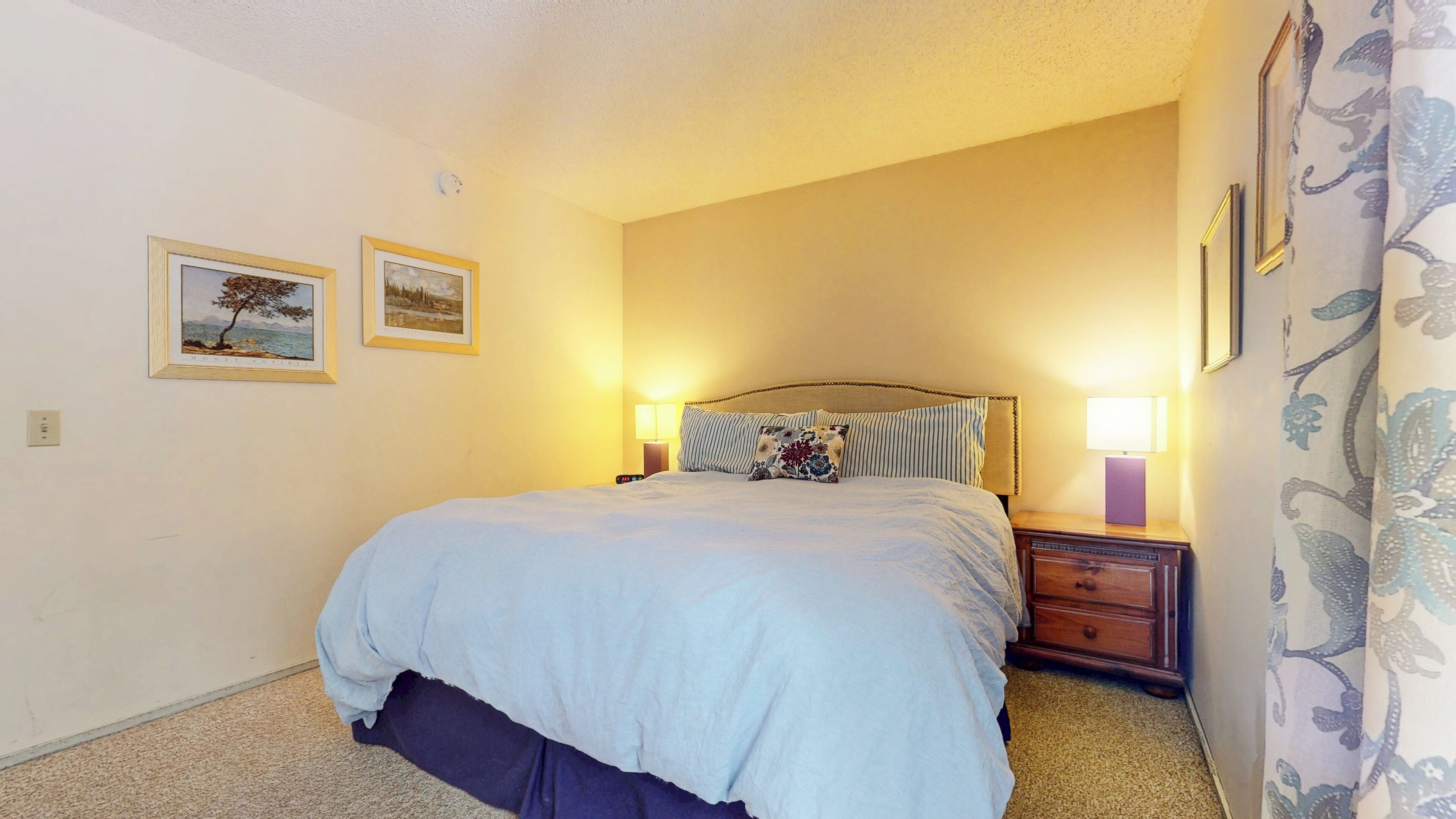 A neatly made bed with a blue comforter and striped pillows, a bedside table with a lamp, and two framed pictures on the wall.