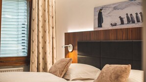 Una habitación de hotel con cama, ventana con persianas, una obra de arte en la pared con pingüinos y cabecera de madera.