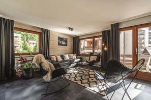 Ein geräumiges Wohnzimmer mit einer Couch, Sesseln, einem gemusterten Teppich und einem Hund.