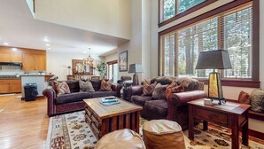 A spacious living room with a dark brown leather sofa, a wooden coffee table, and a patterned rug. There is a dining area with a wooden table and chairs, and a kitchen with wooden cabinets and a black trash bin. - Pet-Friendly, Stunning! On Golf Course! Private Hot Tub, Grill, Washer/Dryer (Mammoth Lakes)
