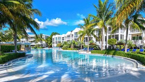 Een zwembad van een resort met palmbomen, een heldere blauwe lucht en witte gebouwen.