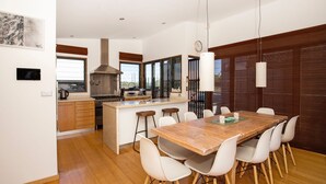 A modern kitchen with a central island, a dining area with a wooden table and white chairs, and a large window providing natural light. - Illaroo - Absolute Beachfront with Spa (Culburra Beach)