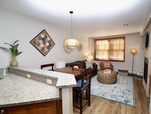 A modern kitchen with granite countertops, a dining area with wooden furniture, a large window with blinds, and a fireplace.