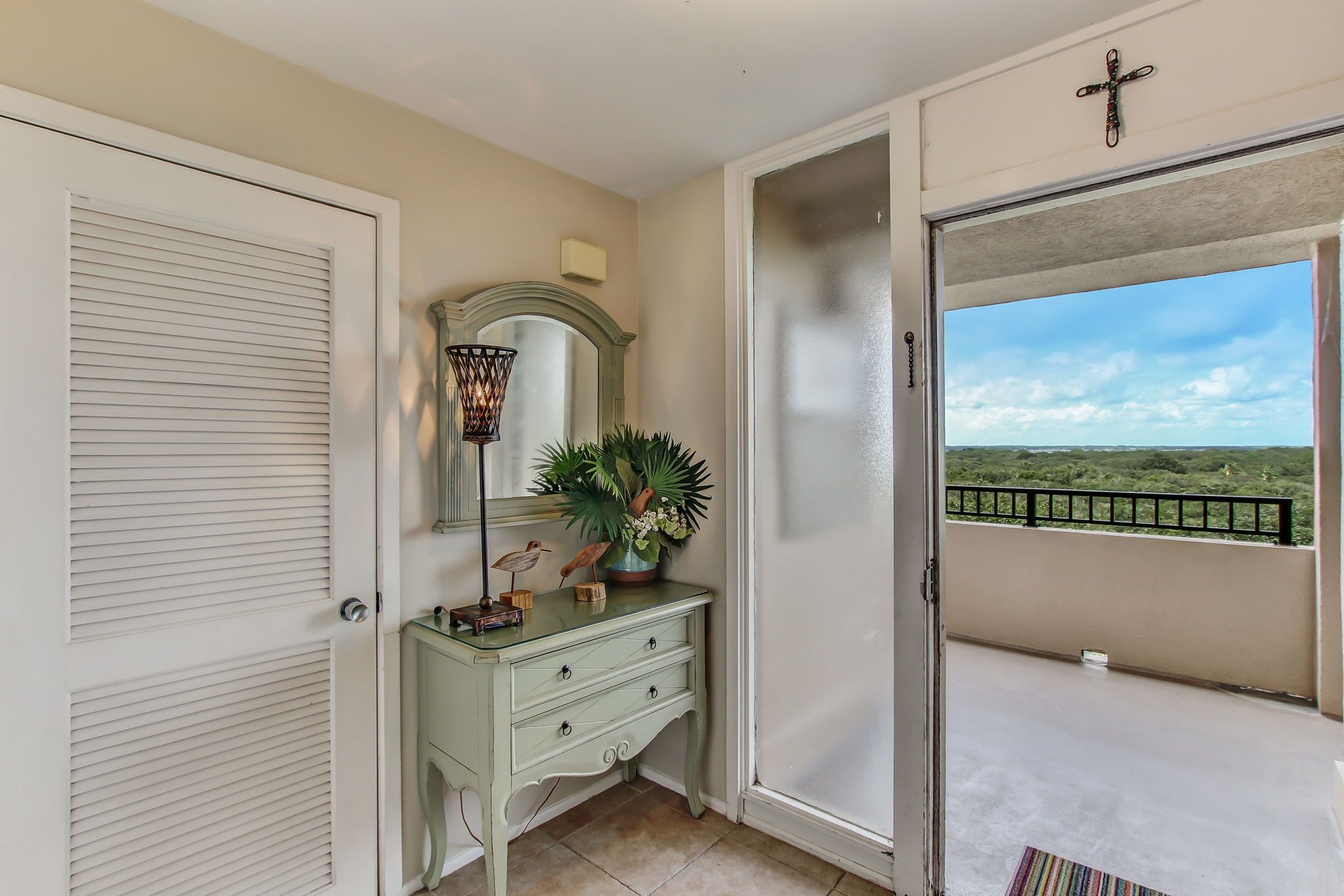 A room with a green vanity, a mirror, a potted plant, and a view of a balcony with a railing.