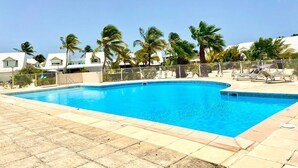 A rectangular swimming pool with clear blue water, surrounded by a tiled deck. Palm trees are visible in the background, and there are lounge chairs and umbrellas on the right side. - Until 6 people, 2 spacious bedrooms, front of the sea, pools tennis parking BBQ (Collectivité de Saint-Martin)