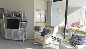 A room with a TV on a white entertainment unit, wicker chairs, a table, and a large window with a view. - One Ocean Place 509 - Oceanfront in Garden City Beach (Garden City)
