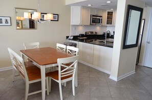 A dining area with a wooden table and white chairs, a kitchen with white cabinets and stainless steel appliances, and a large mirror on the wall. - One Ocean Place 509 - Oceanfront in Garden City Beach (Garden City)