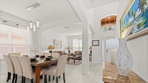A dining area with a wooden table, chairs, and pendant lights. There is a living area with a sofa and a painting on the wall. - 483-Stunning 6-Bed Home w/ Private Pool, Quiet Conservation View (Davenport)