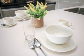 A white dining table with a white bowl, a glass, a spoon, and a fork. There is a potted plant with yellow flowers on the table. - Bell Suites by Salaam Suites (Sepang)