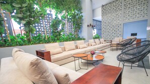 A modern lounge area with a sofa, chairs, and tables, surrounded by greenery and a patterned wall.