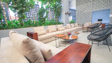 Un espacio lounge moderno con sofá, sillas y mesas, rodeado de follaje y una pared con diseño.