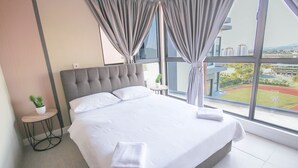 A modern hotel room with a large bed, a small side table with a plant, and a view of a sports field through large windows.