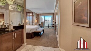 A hotel room with a large bed, a desk, a chair, a sofa, a dining table, a kitchen area, and a balcony with a view.