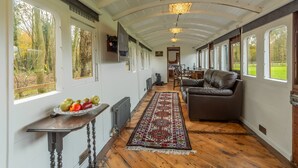 A long, narrow room with wooden floors, a patterned rug, a leather sofa, and a dining table with chairs. - The Railway Carriage, Melton Constable, Norfolk (Melton Constable)