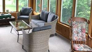A cozy room with a wicker sofa, a glass coffee table, and a wooden chair with floral upholstery. Large windows offer views of greenery outside.