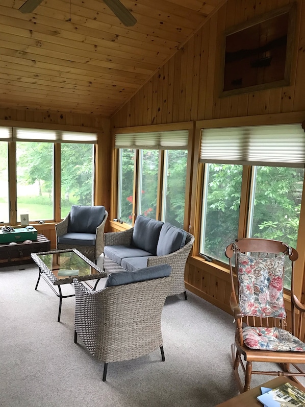 A cozy room with a wicker sofa, a glass coffee table, and a wooden chair with floral upholstery. Large windows offer views of greenery outside.